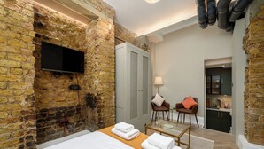A room with a stone wall, a flat-screen TV, a bed with white linens, a glass table, two chairs, and a small kitchenette. - Brixton Lofts - Deluxe Studio (No.2) (London)
