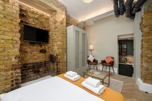 A room with a stone wall, a flat-screen TV, a bed with white linens, a glass table, two chairs, and a small kitchenette. - Brixton Lofts - Deluxe Studio (No.2) (London)