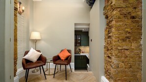 A narrow hallway with a brick wall, two leather chairs with cushions, a small table, a lamp, and a glimpse of a kitchen area. - Brixton Lofts - Deluxe Studio (No.2) (London)