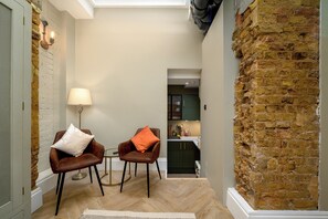 A narrow hallway with a brick wall, two leather chairs with cushions, a small table, a lamp, and a glimpse of a kitchen area. - Brixton Lofts - Deluxe Studio (No.2) (London)