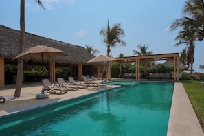 Una piscina con sillas de descanso y sombrillas, rodeada de palmeras y construcciones con techo de paja.