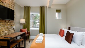 Une chambre d’hôtel avec un mur en briques, un téléviseur à écran plat, un bureau en bois, un lit avec des oreillers blancs et bruns, et une fenêtre avec des rideaux.
