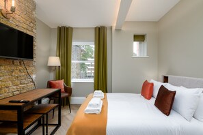 Ein Hotelzimmer mit Backsteinwand, einem Flachbildfernseher, einem Holztisch und einem Bett mit weißen und braunen Kissen sowie einem Fenster mit Vorhängen.