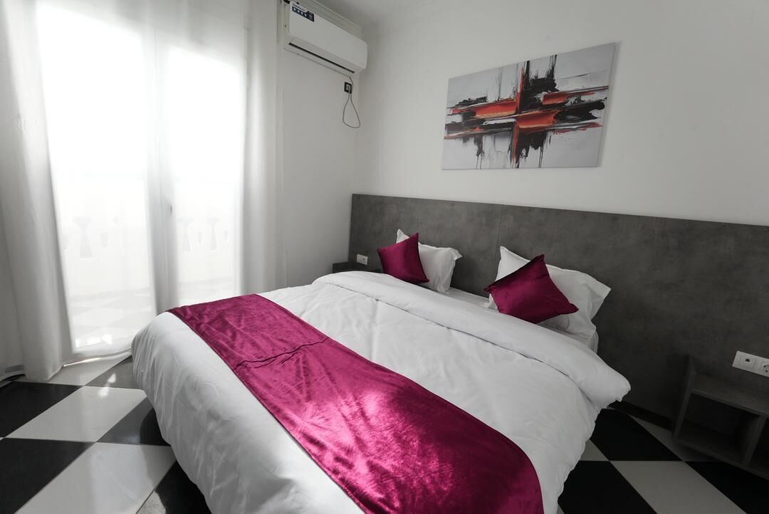 A hotel room with a checkered floor, a bed with white and pink bedding, a grey headboard, and a wall-mounted air conditioner.