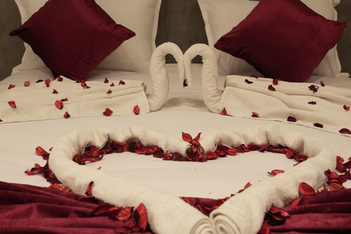 A neatly made bed with a heart-shaped towel arrangement and rose petals, two red pillows, and a gray headboard.