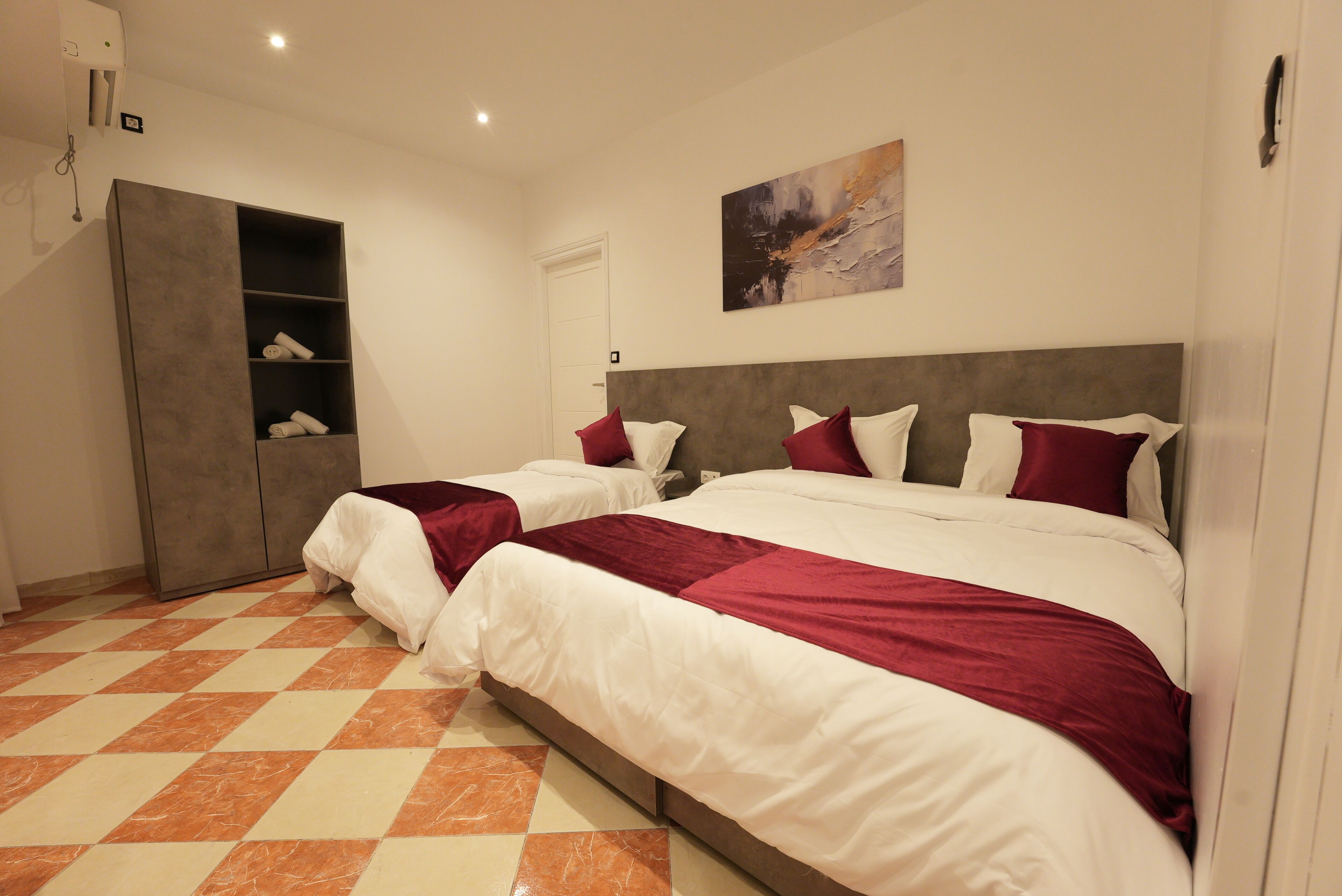 Une chambre d’hôtel avec deux lits, une armoire, un tableau au mur et un sol à carreaux.