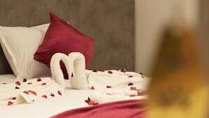 Una cama con ropa de cama blanca y roja, cabecera gris y pétalos de rosa esparcidos sobre la cama.
