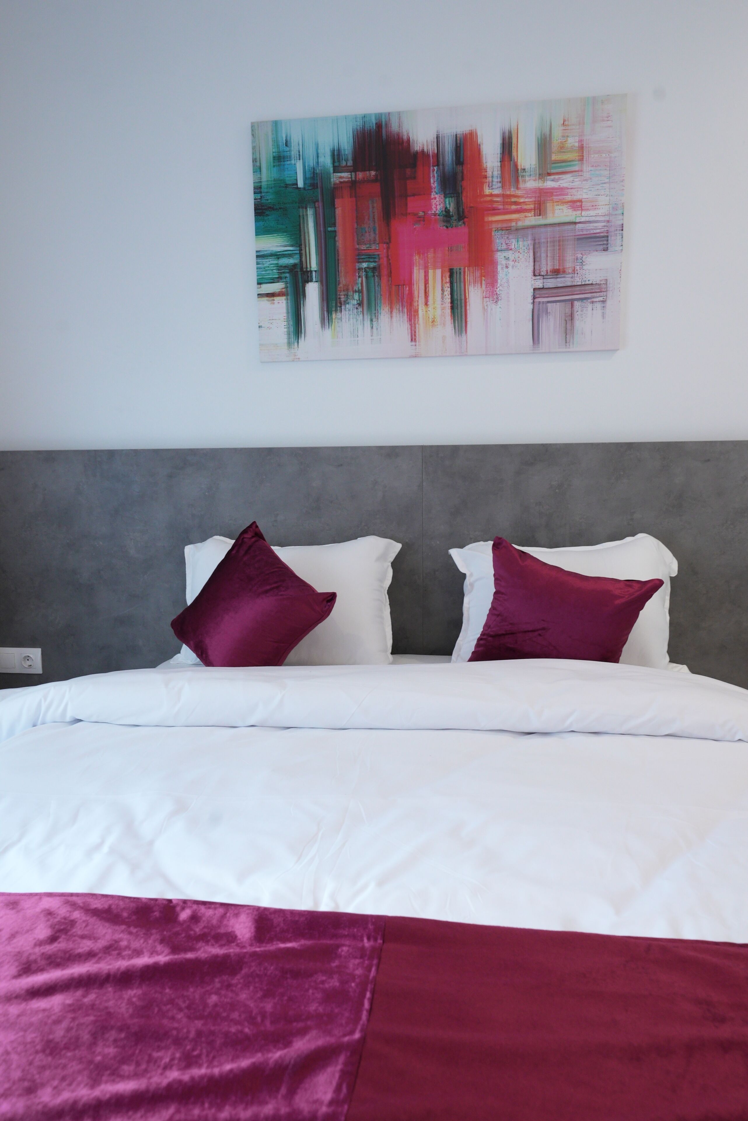 Ein Bett mit weißer Bettwäsche, zwei dunkelroten Kissen, einem grauen Kopfteil und einem farbenfrohen, abstrakten Gemälde an der Wand.