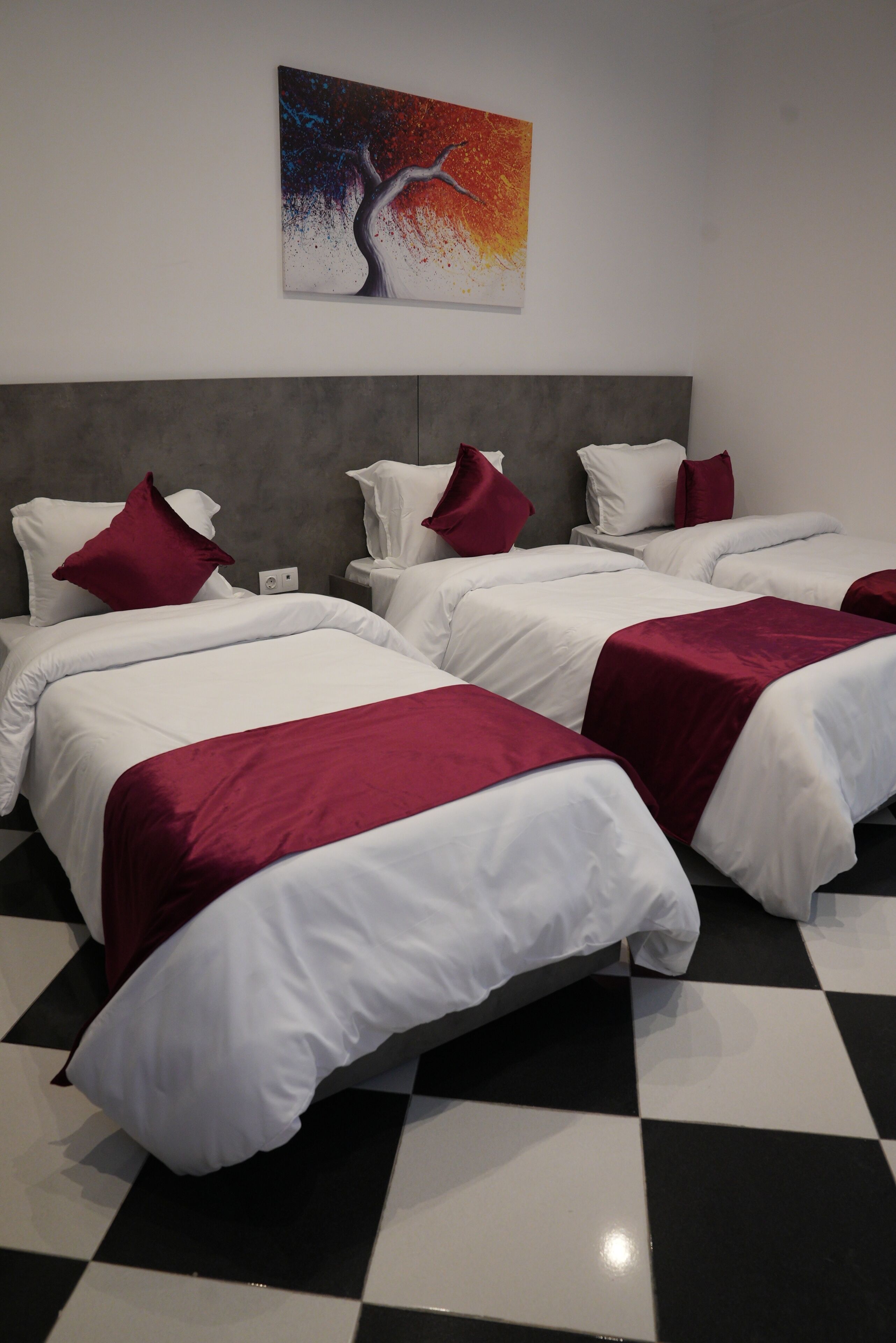 A hotel room with four beds, each with white bedding and red accents, a grey headboard, and a checkered floor.