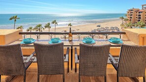 Ein Balkon mit Korbstühlen und blauen Tellern, mit Blick auf einen Strand mit Palmen und einem Hotel im Hintergrund.
