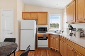 A compact kitchen with wooden cabinets, a white refrigerator, a microwave, a sink, and a dining area with a table and chairs. - Cottage #2 at The Fairway Farm | Disc Golf Getaway (Quinton)