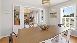 Un comedor con una mesa de madera, sillas blancas, un frutero, una lámpara colgante y vistas a través de una ventana.