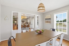 A dining area with a wooden table, white chairs, a fruit bowl, a hanging light fixture, and a view through a window. - Fairway Farmhouse | Stay & Play Disc Golf Course! (Quinton)