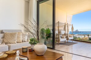 Ein modernes Wohnzimmer mit einer Couch, einer großen Vase mit Zweigen, einem Couchtisch und Blick auf einen Balkon mit Gartenmöbeln.
