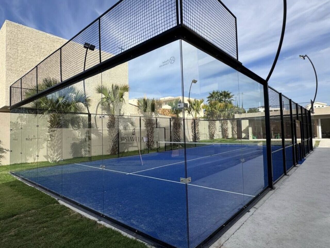 Ein Tennisplatz im Freien mit Glasumrandung, umgeben von einer niedrigen Mauer und Palmen.
