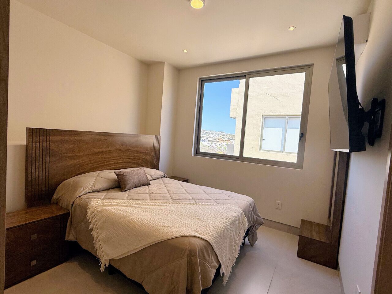 Un dormitorio con un ventanal grande, una cama con cabecera de madera, un televisor montado en la pared y una mesita de noche con cajones.