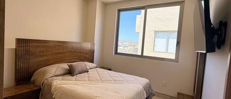 Un dormitorio con un ventanal grande, una cama con cabecera de madera, un televisor montado en la pared y una mesita de noche con cajones.