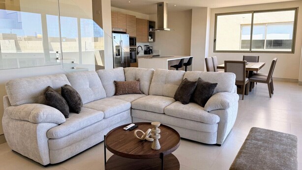 A modern living room with a sectional sofa, a coffee table, and a dining area with chairs and a table. - Penthouse | Ocean Views, Pools & Padel Court (Cabo San Lucas)