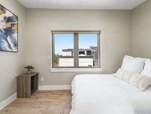 A bedroom with a large window, a bed with white bedding, a small wooden side table with a plant, and a painting on the wall.