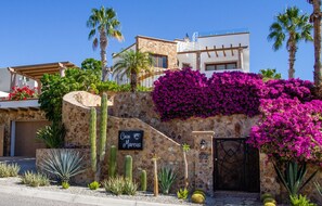 Ein modernes Haus mit Naturstein-Fassade, einem Schild mit der Aufschrift 'Casa Marcus' sowie verschiedenen Kakteen und Bougainvillea-Pflanzen.