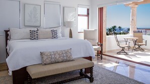 Quarto com cama grande, um banco, uma cadeira e uma mesa numa varanda com vista.