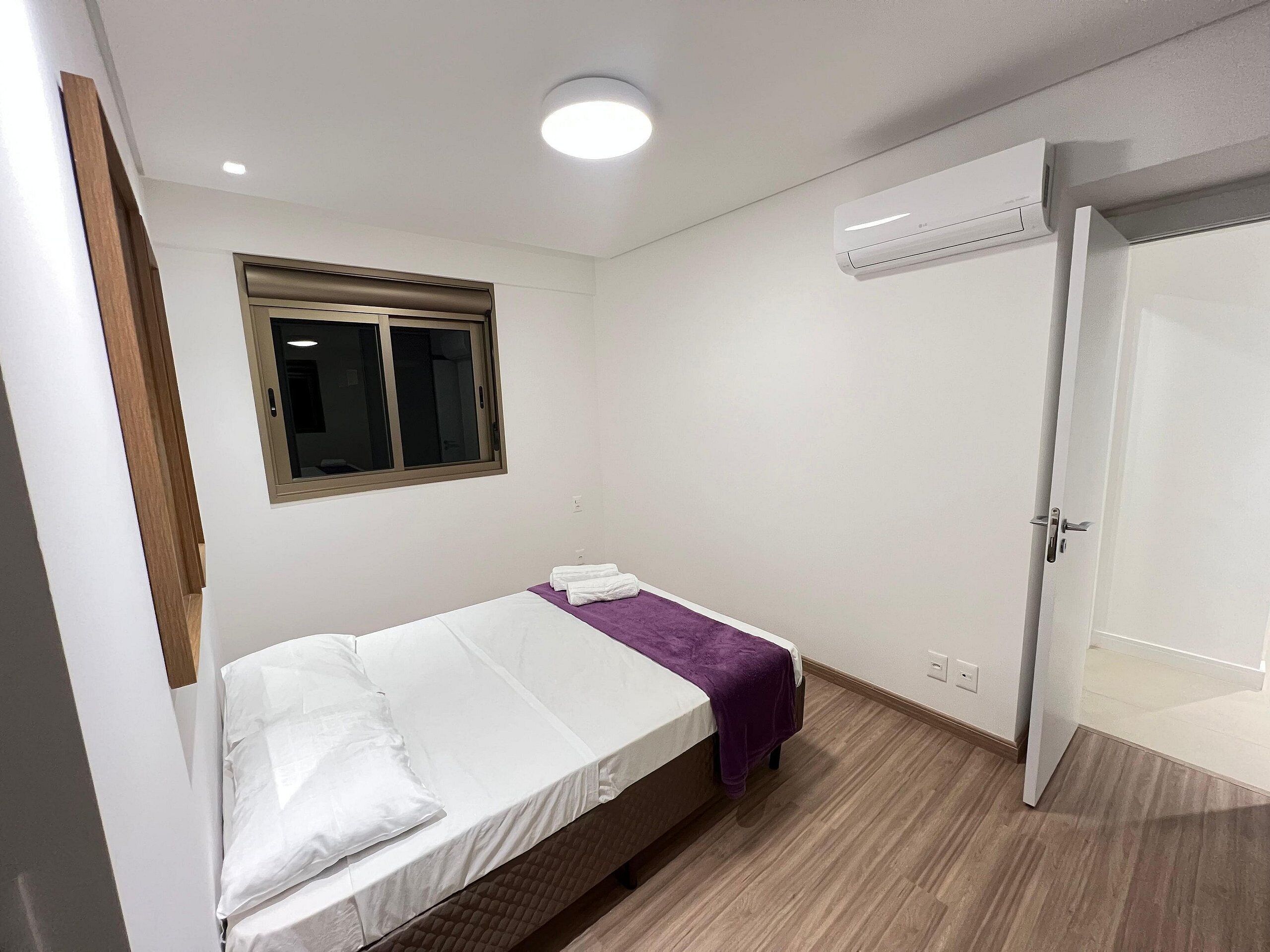 A small, clean room with a bed, a window, an air conditioning unit, and a door.