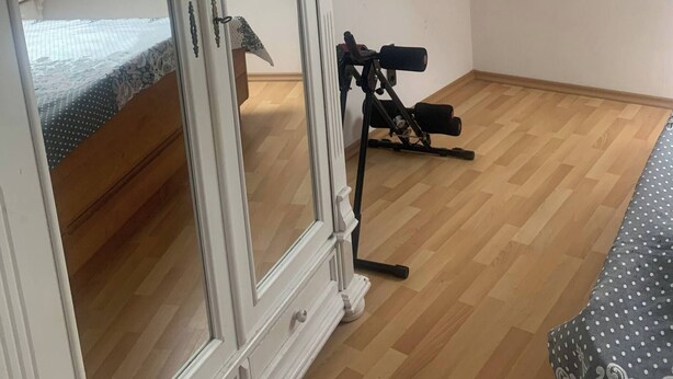 Una habitación con piso de madera, una cama con colcha estampada, un armario blanco y un trípode con una cámara.