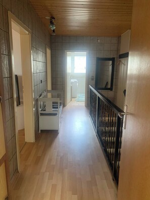 A hallway with a wooden floor, a white bench, a black railing, and a door leading to a bathroom. - Pure Relaxation Quiet and Cozy (Weyhausen)