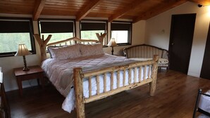 Een bed met houten frame in een kamer met schuine wand, een stoel, een nachtkastje en een lamp.