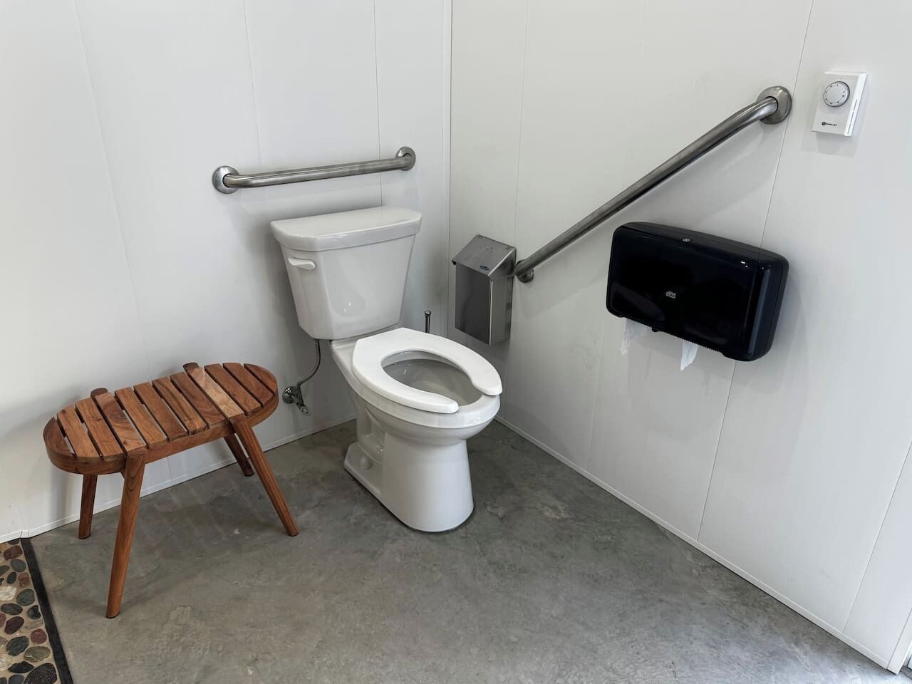 Une salle de bain équipée de toilettes, d’un banc en bois, d’un distributeur de gel hydroalcoolique et d’une barre d’appui.