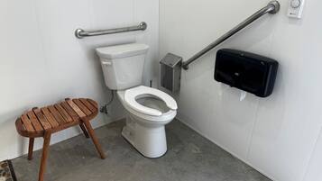Une salle de bain équipée de toilettes, d’un banc en bois, d’un distributeur de gel hydroalcoolique et d’une barre d’appui.