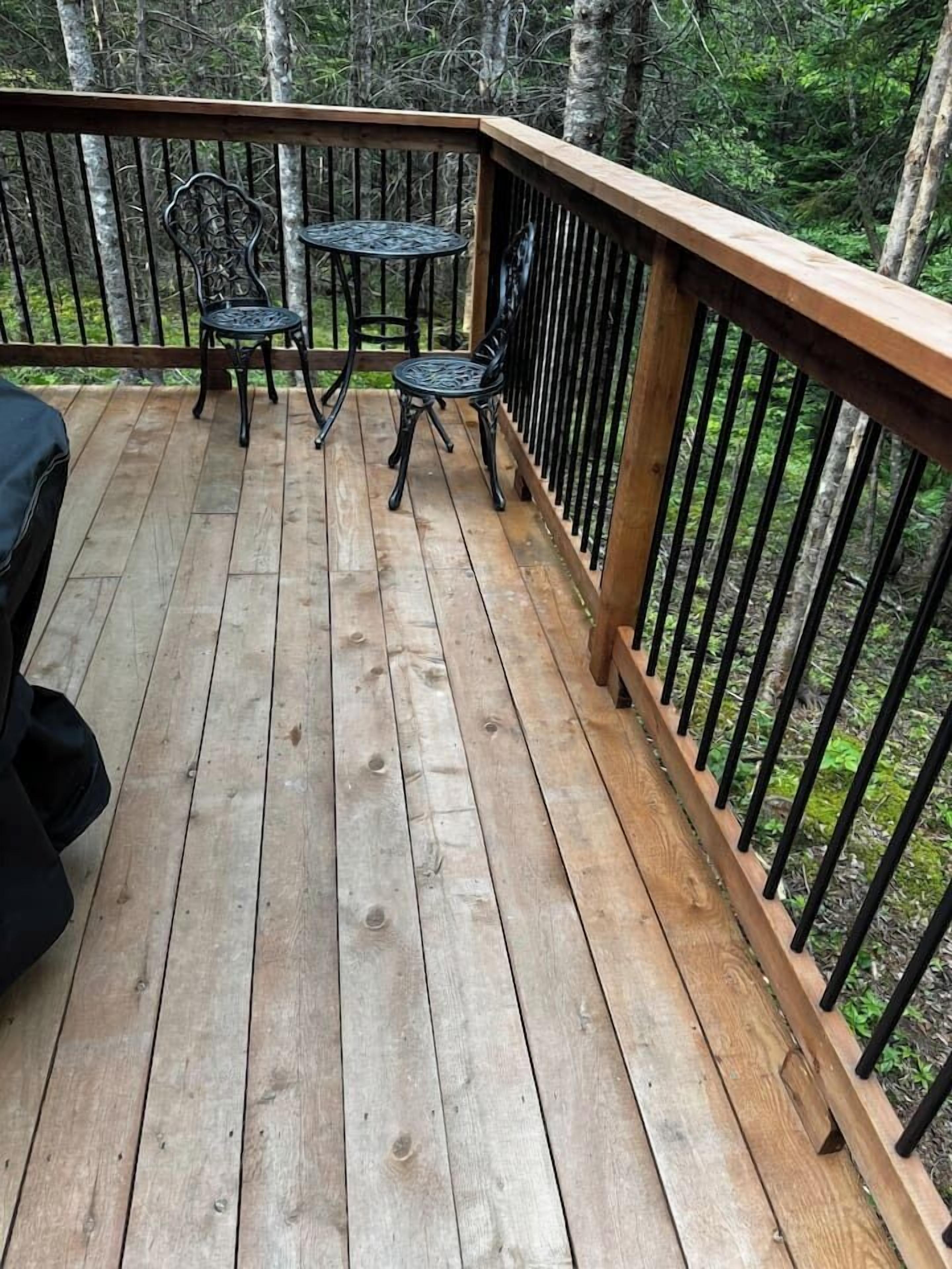 Una terraza de madera con barandillas de metal, dos mesas metálicas y sillas.