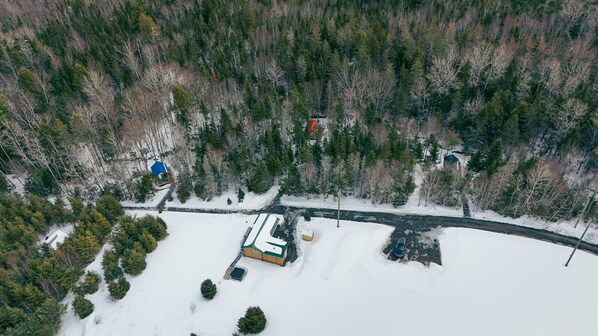 An aerial view of a snowy landscape with a road, a small building with a blue roof, and a forested area.