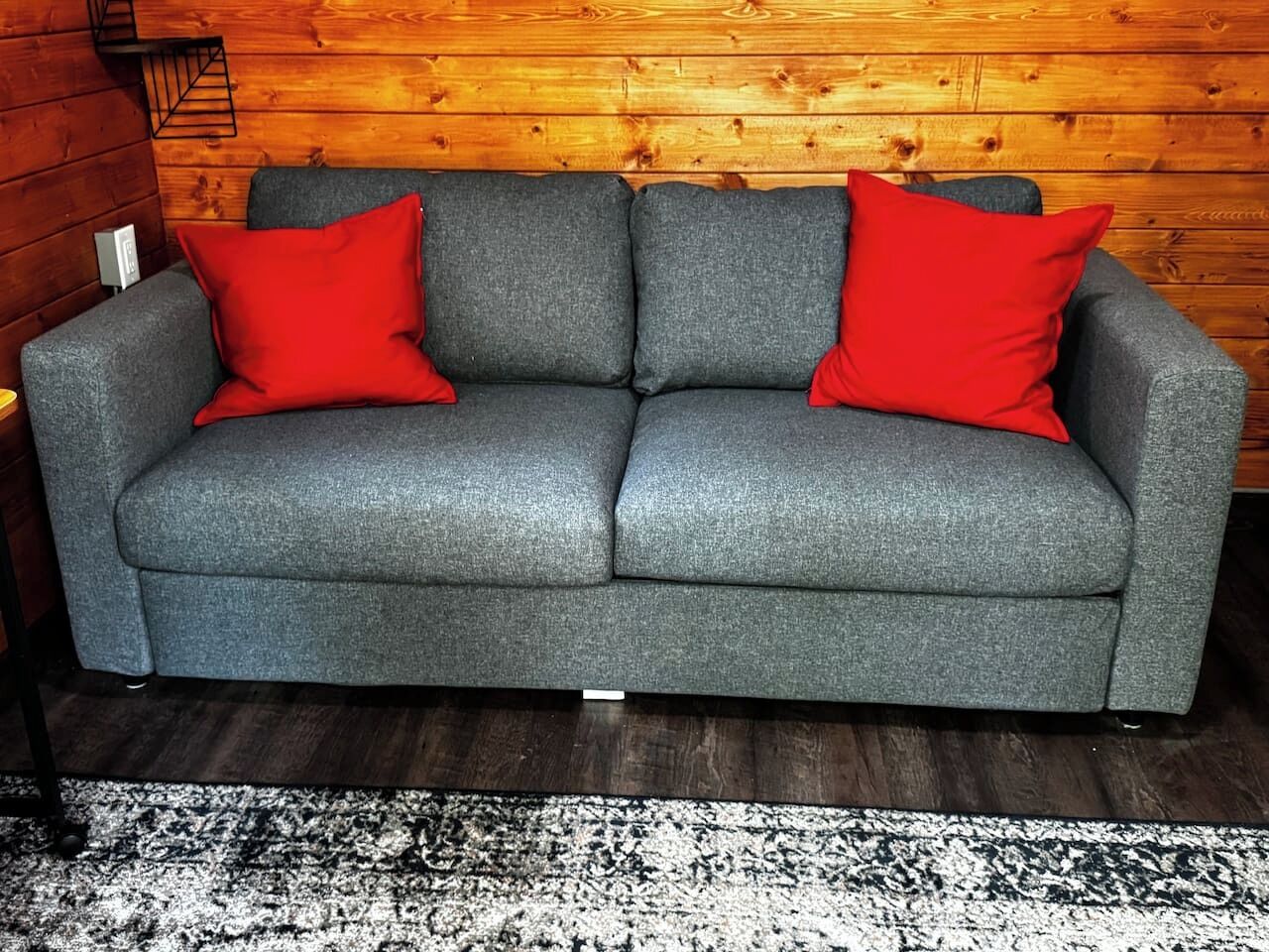 Un canapé gris avec des coussins rouges, adossé à un mur en bois, un tapis noir et blanc et un présentoir en métal noir.