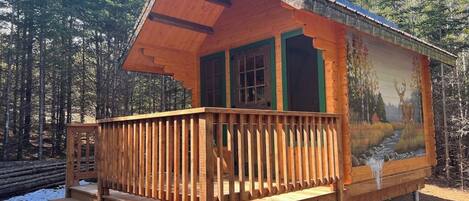 A wooden cabin with a balcony, a painting of a landscape on the side, and a set of stairs leading to the entrance.