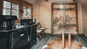 A rustic cabin interior with a kitchenette, a dining table, and a leather rug. - Pet-Friendly Cabin | Sleeps 2 | Private Fire Pit (Mount Uniacke)