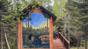A wooden cabin with a glass wall reflecting a forest scene, a deck, and a small outdoor grill. - Private 4 Cabin Retreat | Hot Tub | Tiny Cabins (Mount Uniacke)