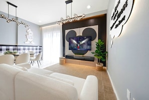 Ein modernes Wohnzimmer mit Flachbildfernseher, weißen Sofas, einem Essbereich mit kariertem Tischläufer und einem dekorativen Wandbild.