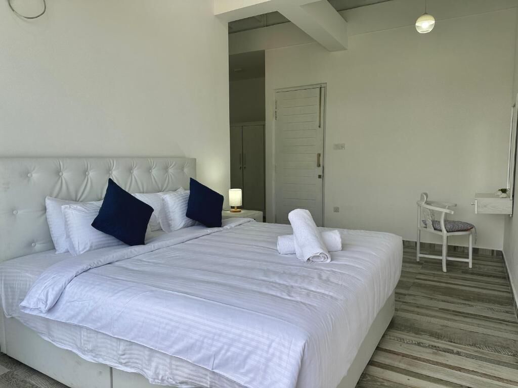 Un lit bien fait, avec du linge de lit blanc et des oreillers foncés, une table de chevet avec une lampe, une chaise et une porte donnant sur une autre pièce.