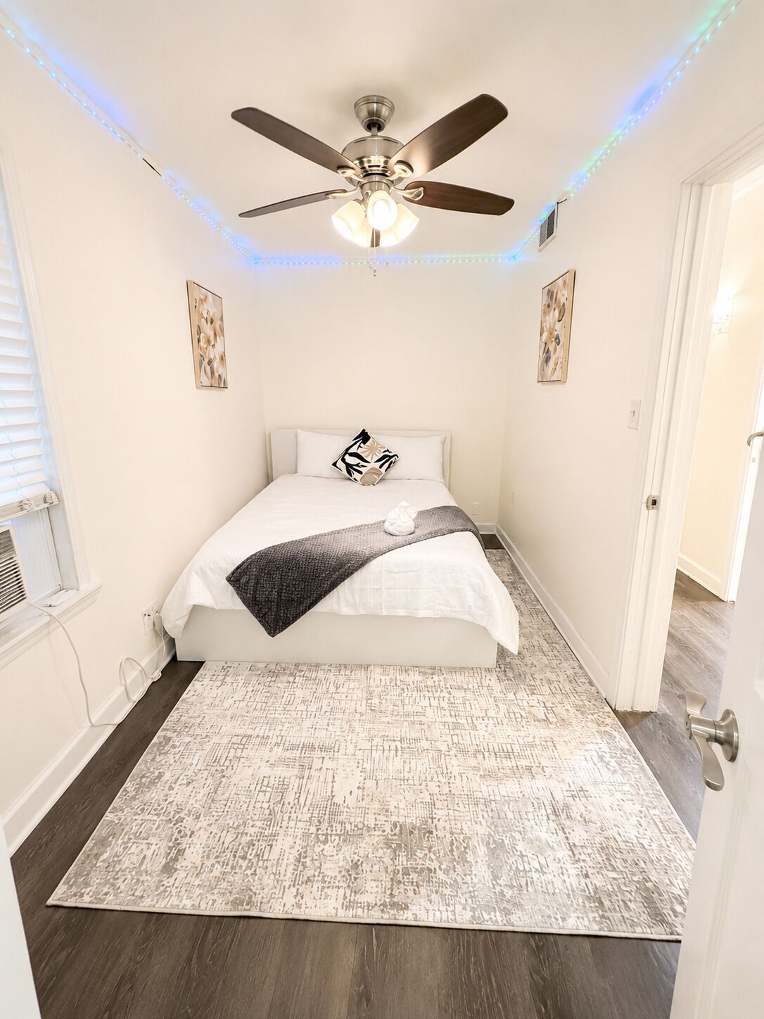 A bedroom with a ceiling fan, a bed with a white comforter, a patterned rug, and artwork on the walls.
