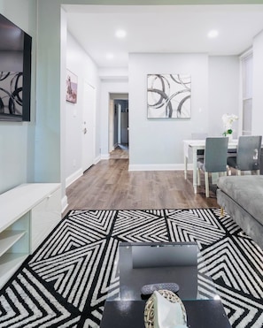 A modern living room with a black and white patterned rug, a flat-screen TV, a grey sofa, and a dining area with a white table and chairs.