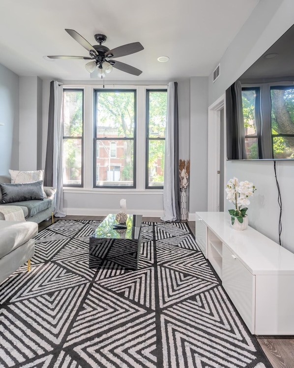 A modern living room with a geometric patterned rug, a ceiling fan, a sofa, a coffee table, and a TV on a stand.
