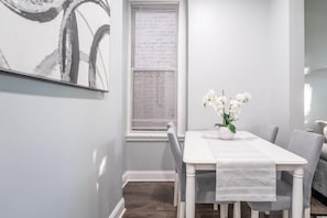 A dining area with a white table, chairs, a vase with flowers, and a window with blinds.