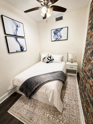 Quarto com cama, mesa de cabeceira, luminária, ventoinha no teto, obra de arte e tapete estampado.