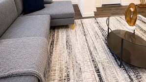 Ein modernes Wohnzimmer mit einem grauen Ecksofa, einem runden Glastisch und einem Teppich mit Muster. Im Hintergrund sind Einbauschränke mit Pflanzen und eine weiße Tür zu sehen.