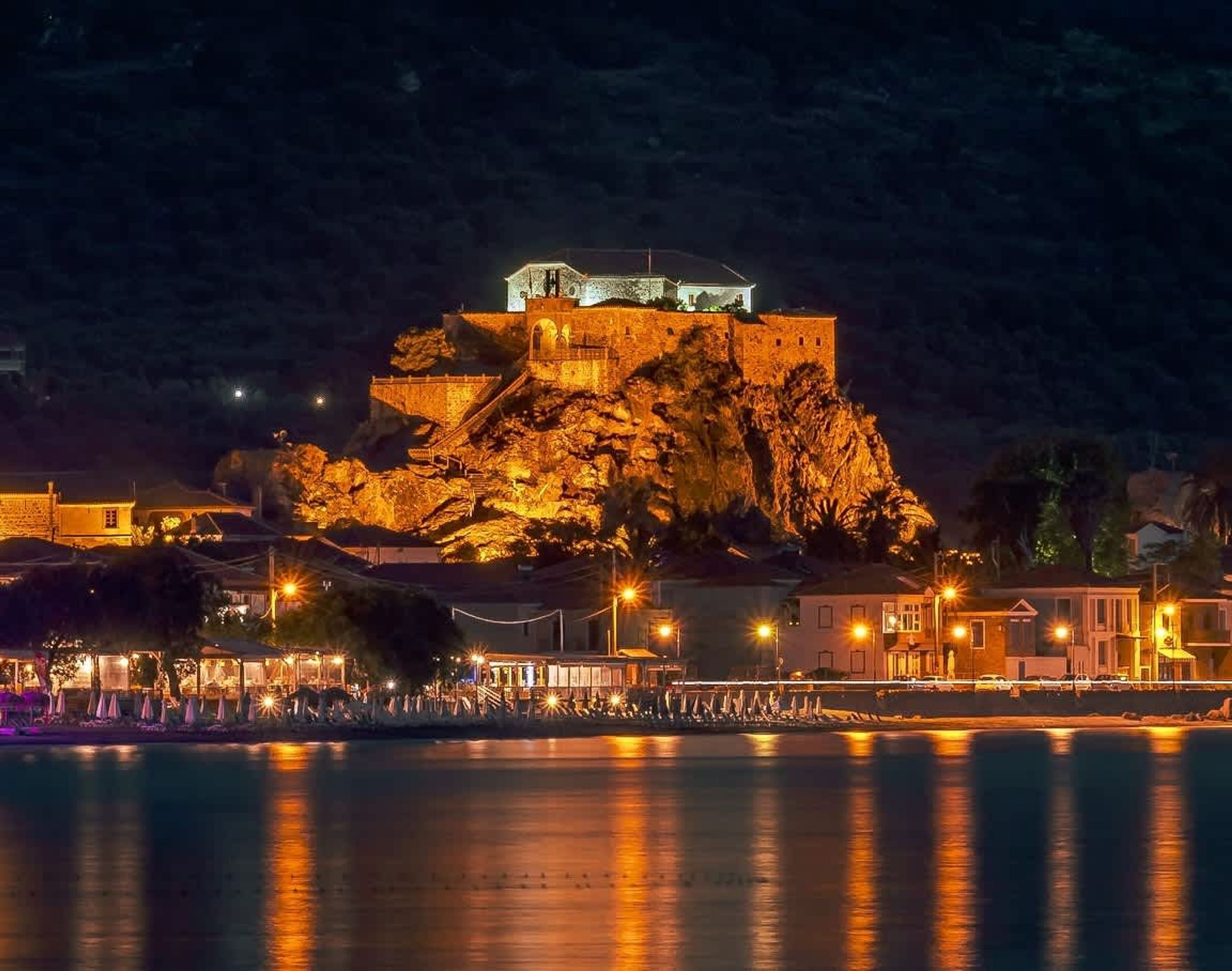 A historic stone fortress on a cliff overlooking a calm body of water, with a well-lit promenade and buildings in the foreground.