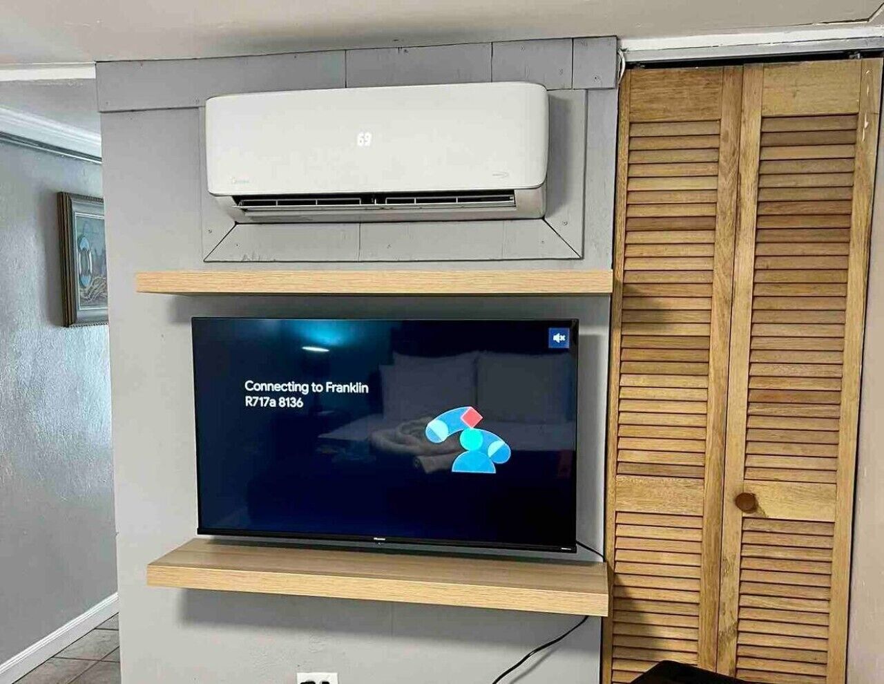A room with a mounted air conditioner, a flat-screen TV displaying a connection message, and a wooden wardrobe.