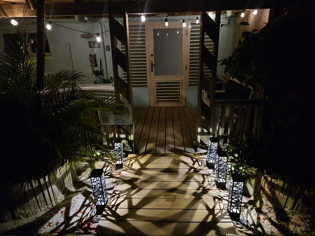 A wooden pathway leading to a door with a glass panel, surrounded by potted plants and string lights.