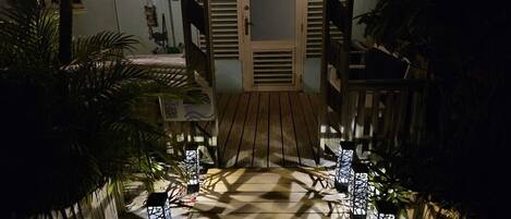 A wooden pathway leading to a door with a glass panel, surrounded by potted plants and string lights.