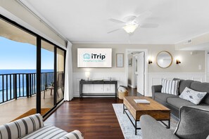 Ein geräumiges Wohnzimmer mit großem Fenster zum Meer, Deckenventilator, Couchtisch aus Holz, graues Sofa und ein Fernseher, der das Logo von iTrip Panama City Beach zeigt.